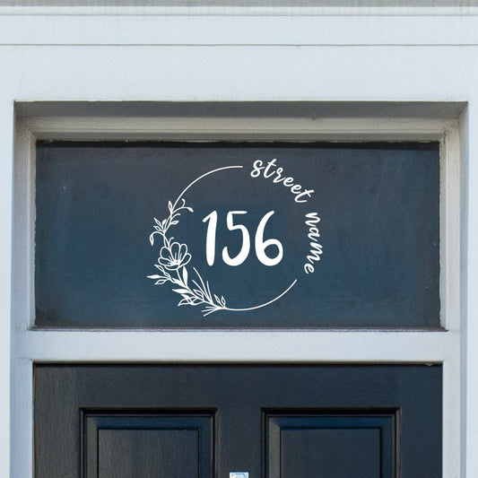 A house door number and street name with floral decorations vinyl decal sticker stuck on to the window above a black door.